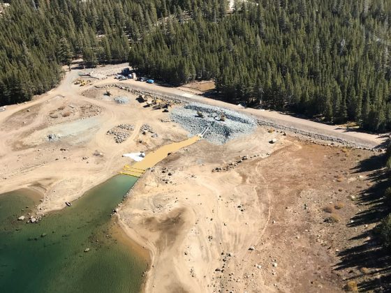 Aerial view of rock-fill buttress on upstream side of embankment dam in a mountainous setting.