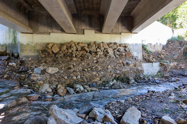 Under a bridge, there is a stream flowing with a natural rock and dirt streambed. The bridge foundation is marked with scour.