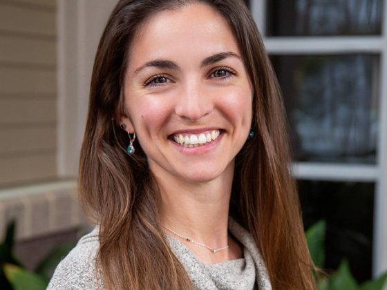 A woman with long brown hair smiles for a headshot in front of a house.