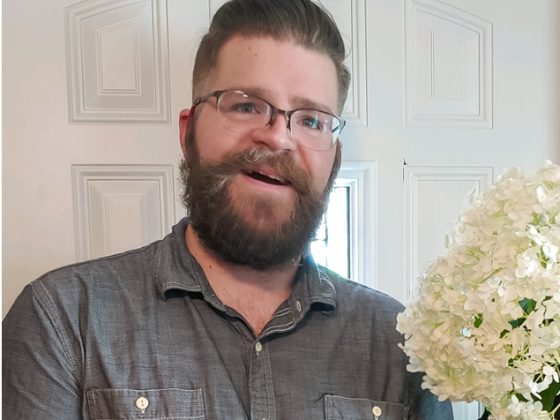 Jesse is standing in his doorway holding a bunch of white flowers he picked from his garden. He is wearing a gray shirt and black gloves.