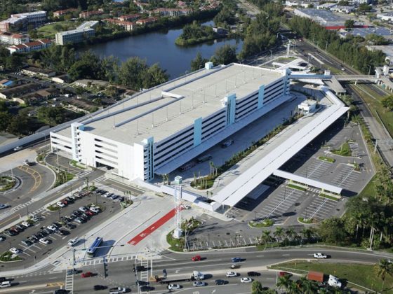 Arial view of a white parking garage with cars and busses driving and water at the top left.