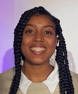 A woman with braids wearing a white shirt with a tan sweater over it.