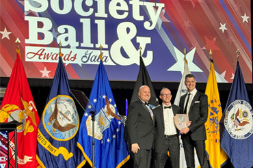 The image depicts three men standing on a stage during an awards gala. They are all dressed in formal attire, with two of them holding a plaque between them. Behind them is a large screen displaying the text "2024 Society Ball & Awards Gala." Several flags, including those representing different branches of the U.S. military, are arranged in the background.