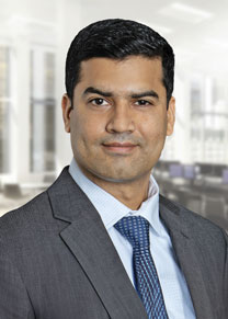 A man is posing for his headshot. He wears a blue checked shirt, dark blue patterned tie, and gray suit jacket.