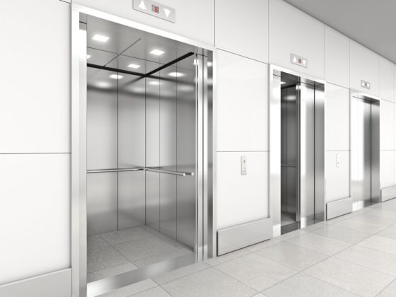 Three shiny metallic elevators in a row along a wall. One elevator door is open, one is partially open, and one is closed.